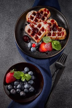 Delicious waffles heart shape with  powdered sugar and berries on ceramic black plate and saucer with berries on grey background. Top view. Sweet meal. Dessert. Serving food