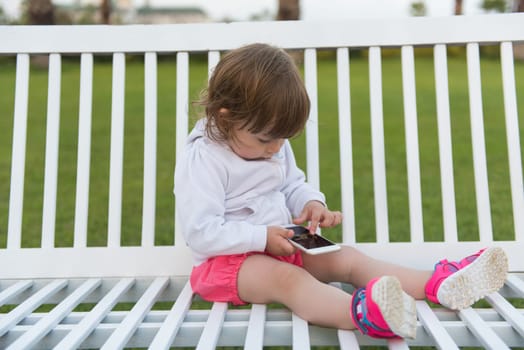 beautiful little girl playing with mobile phone while sitting on the bench in the park
