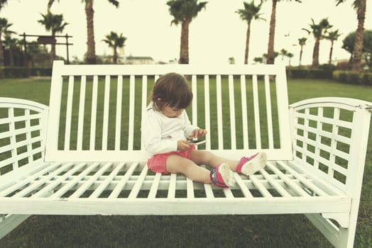 beautiful little girl playing with mobile phone while sitting on the bench in the park