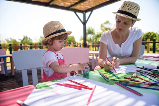 young mother and her little daughter cheerfully spending time together using pencil crayons while drawing a colorful pictures in the outside playschool