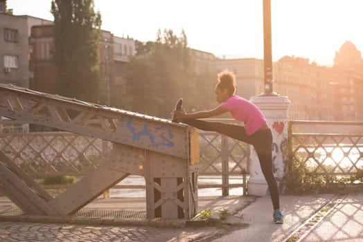 young beautiful African American woman doing warming up and stretching with her leg raised to the bridge before the morning run with the sunrise in the background