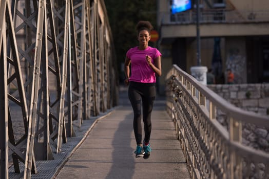 Young sporty african american woman running on sidewalk across the bridge at early morning jogging with city sunrise scene in background