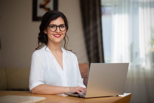 Happy businesswoman with glasses working from home. Smiling woman with white shirt typing on laptop in homeoffice. Positive female employee smiling to the camera.