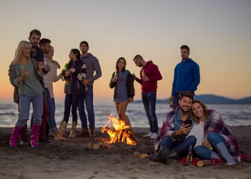 Couple using cell phone during beach party with friends drinking beer and having fun colored filter
