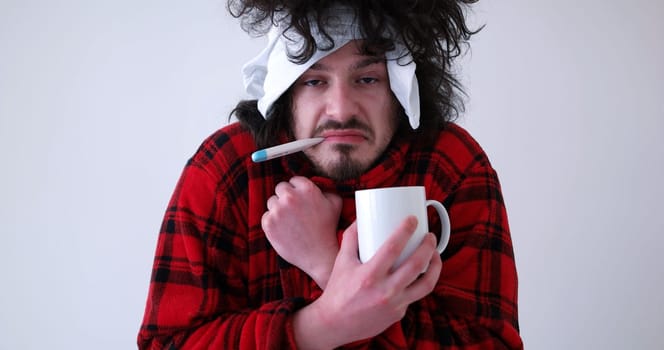 young Man with flu and fever wrapped holding cup of healing tea isolated over white