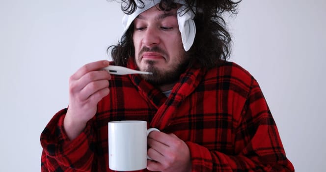 young Man with flu and fever wrapped holding cup of healing tea isolated over white