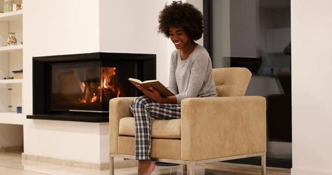 Young beautiful african american woman sitting in front of fireplace at home on a cold autumn day and reading book