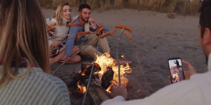 Group of young friends sitting by the fire late at night, grilling sausages and drinking beer, talking and having fun