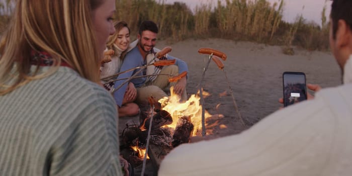 Group of young friends sitting by the fire late at night, grilling sausages and drinking beer, talking and having fun