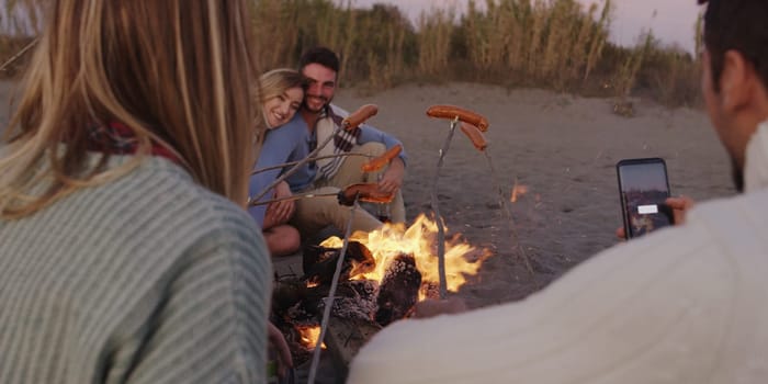 Group of young friends sitting by the fire late at night, grilling sausages and drinking beer, talking and having fun