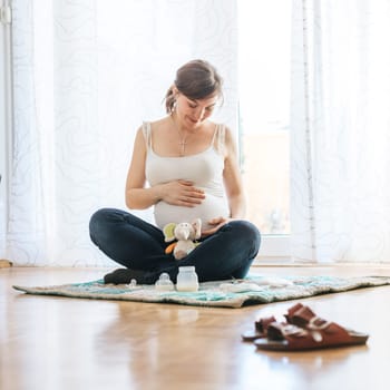 Happy Caucasian pregnant mother is sitting on the floor, touching her tummy, blue jeans