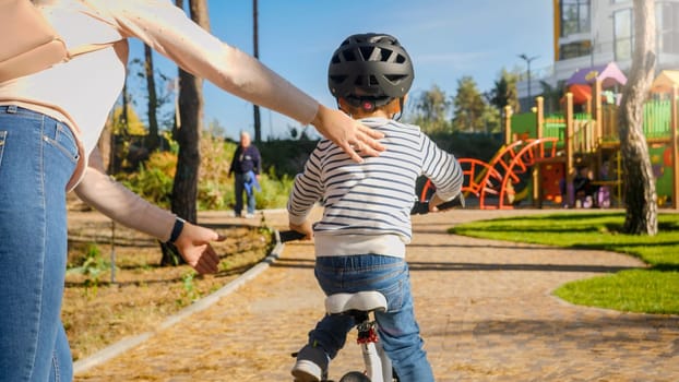 Young moter holding and supporting her little son riding bicycle for first time at park.