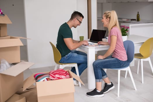 Young couple moving in a new home. Man and woman at the table using notebook laptop computer and plans with boxes around them