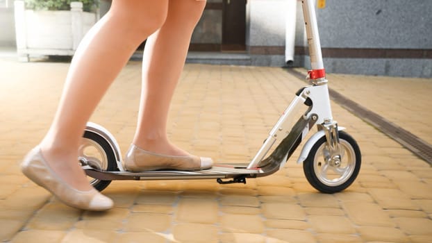 Closeup photo of young woman in ballet flats riding fast on the scooter on city street.