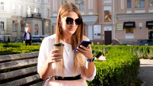 Portrait of young woman in sunglasses sitting on the bench at park with coffee paper cup and typing message on smartphone.