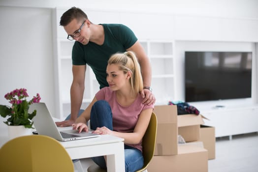 Young couple moving in a new home. Man and woman at the table using notebook laptop computer and plans with boxes around them
