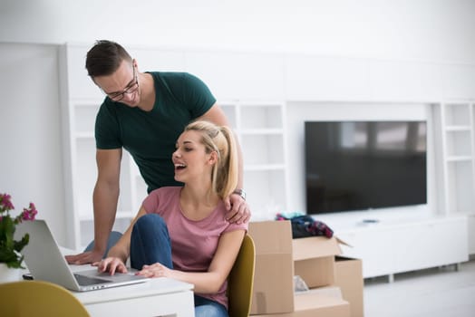 Young couple moving in a new home. Man and woman at the table using notebook laptop computer and plans with boxes around them