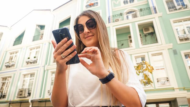 Portrait of beautiful young woman in sunglasses typing message on smartphone on city street.