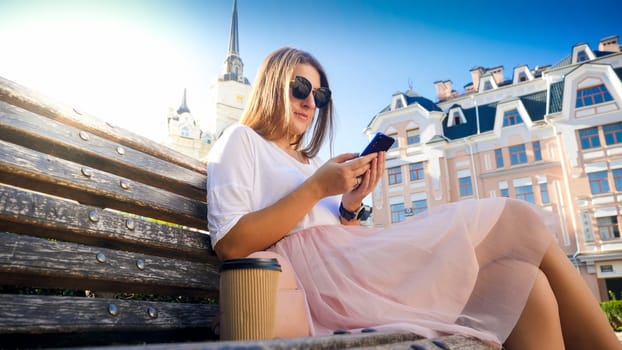 Portrait of beautfiul woman with long hair sitting on the bench and typing message on mobile phone.