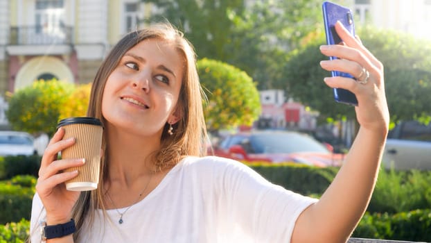 Portrait of smiling woman with coffee cup making selfie photo on smartphone at park.