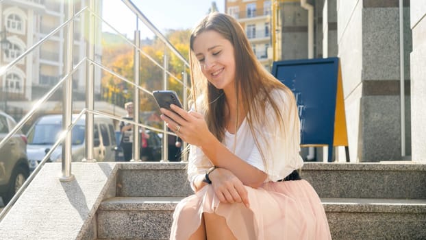 Toned photo of happy smiling girl sitting on stairs on street and browsing internet on smartphone.