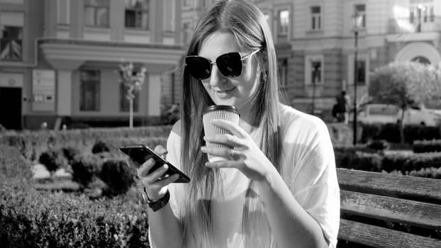 Black and white portrait of smiling girl drinking coffee and browsing internet on smartphone while sitting on the bench at park.