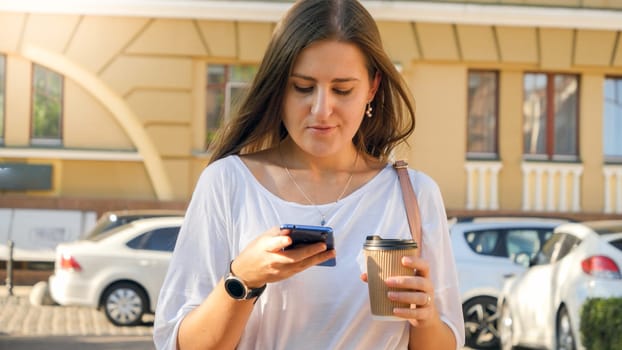 Portrait of beautiful female worker walking on city street with coffe cup and browsing smartphone.
