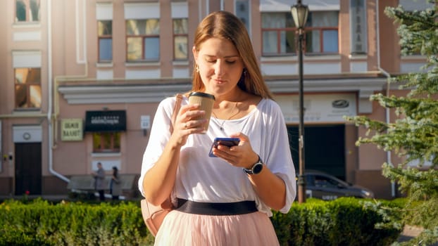 Beautiful young woman holding coffee paper cup in one hand and smartphone in other hand.