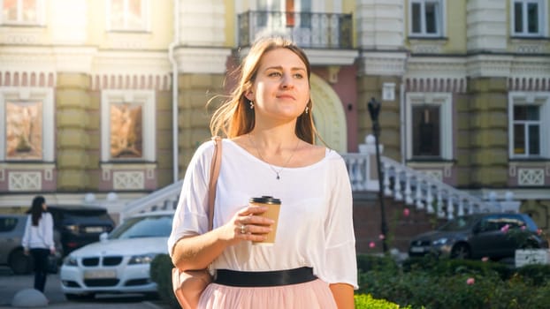 Portrait of stylish young woman walking on city street with coffee-to-go paper cup.