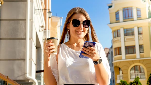Portrait of beautiful smiling young woman holding cup of coffee walking on street and using smartphone.