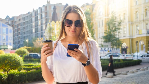 Portrait of beautiful young woman long hair holding coffee cup walking on street and typing message in social media on smartphone.