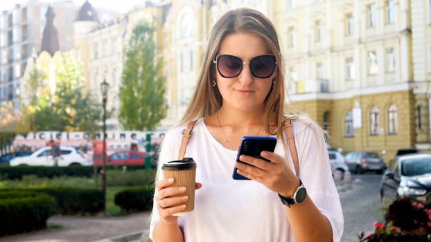 Portrait of beautiful female student with cup of coffee browsing internet on smartphone.