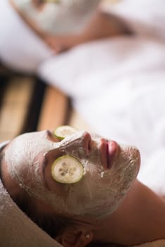Beautiful young woman is getting facial clay mask at spa, lying with cucumbers on eyes