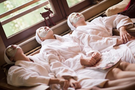 beautiful young women  getting facial clay mask at spa, lying with cucumbers on eyes