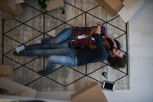 Top view of attractive young multiethnic couple moving, holding hands, looking at camera and smiling while lying among cardboard boxes