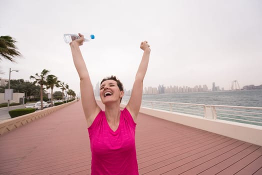 young woman celebrating a successful training run on the promenade by the sea with a bottle of water and her hands raised in the air with a big city in the background