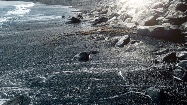 Seascape of bright sun shining on black volcanic sea beach and blue ocean waves.