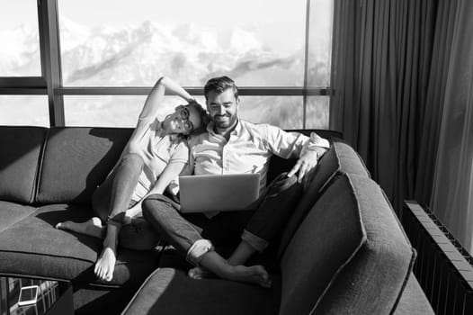 Young couple relaxing at  home using laptop computers reading in the living room near the window on the sofa couch.