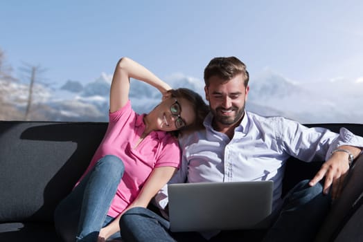 Young couple relaxing at  home using laptop computers reading in the living room near the window on the sofa couch.