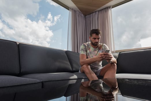 young man sitting on sofa and using a mobile phone  near the window at home