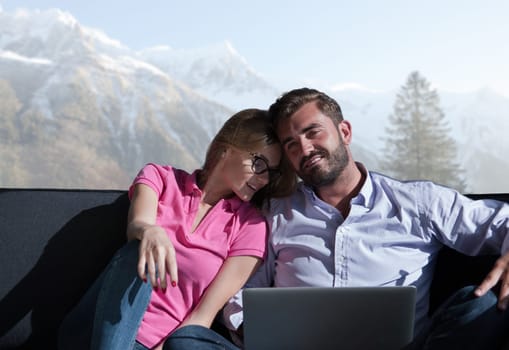Young couple relaxing at  home using laptop computers reading in the living room near the window on the sofa couch.