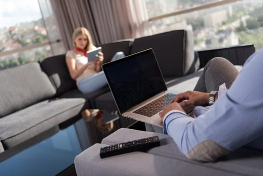 Young couple relaxing at home using tablet and laptop computers reading near the window on the sofa couch.