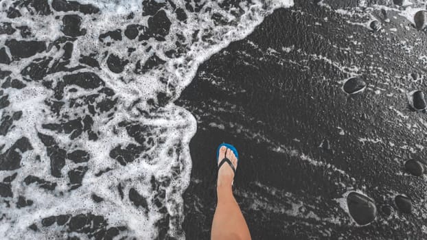 View from the top on male feet in flip flops walking on black volcanic sandy sea beach.