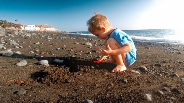Little toddler boy playing on the sea beach with plastic shovel and building sand castle.