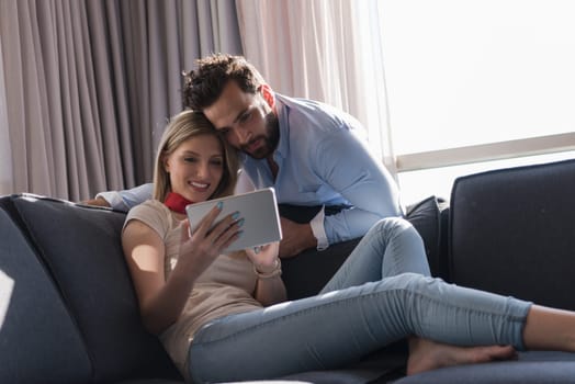 Young couple relaxing at  home using tablet computer reading in the living room near the window on the sofa couch.