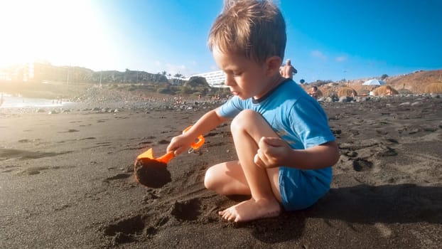 Portrait of little toddler boy digging wet sand with toy shoel on sea beach.