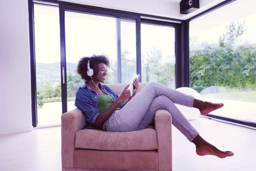 young beautiful African American woman enjoys listening to music with headphones and tablet in your armchair in her luxury home