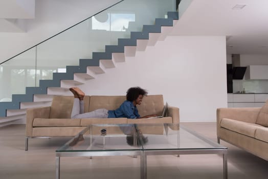 Smiling young African American woman using laptop while relaxing on sofa at luxury home