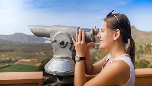 Portrait of happy smiling female tourist looking on ountains through coin operated binoculars.