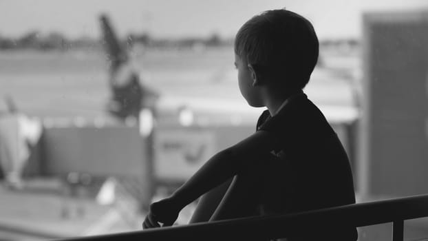 Black and white upset little boy sitting in airport terminal and looking on big airplanes.